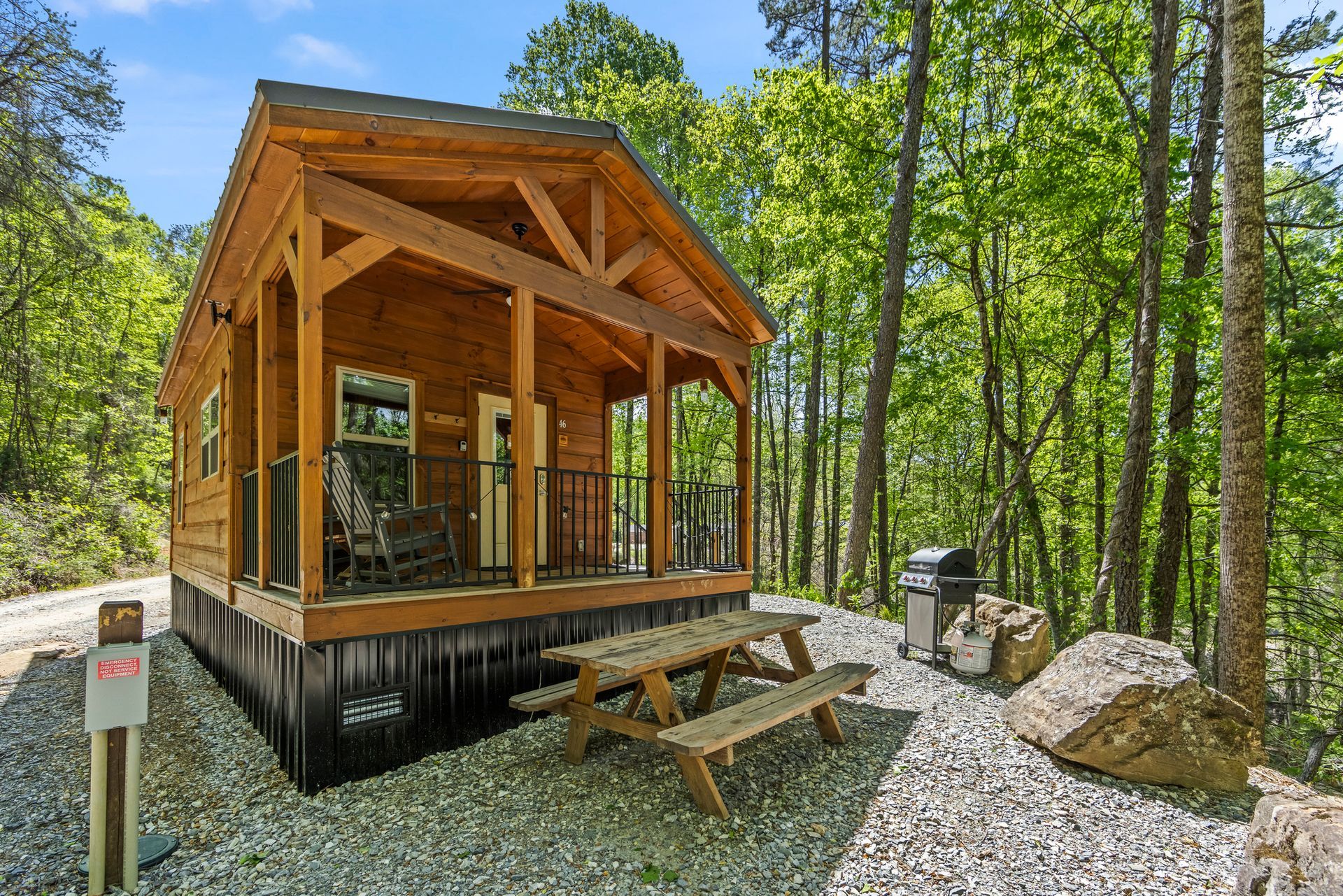 Wooden cabin on a gravel lot surrounded by trees, with a picnic table and large rocks nearby.