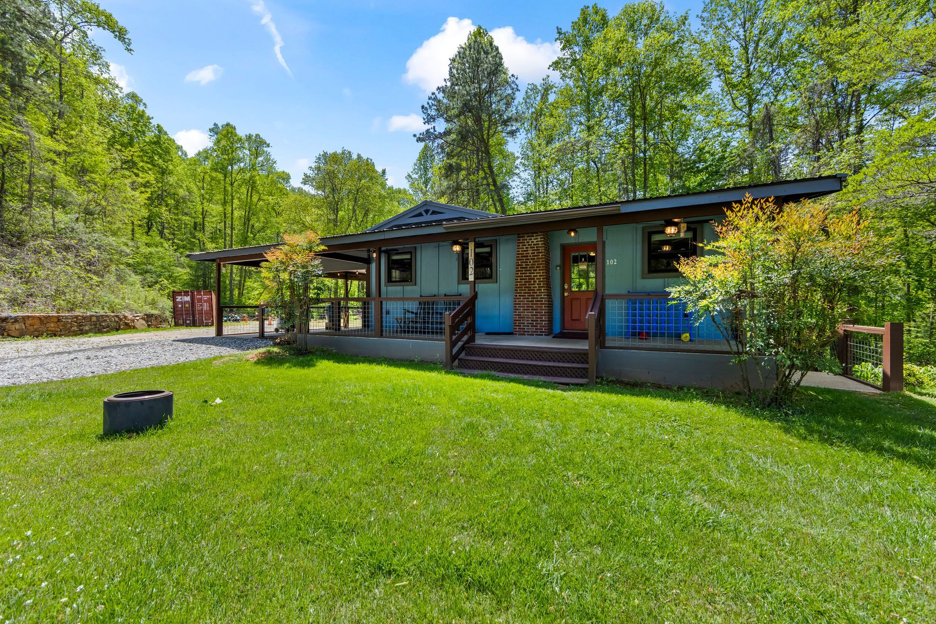 Modern cabin with blue siding and red door on a green lawn, surrounded by trees under a clear sky