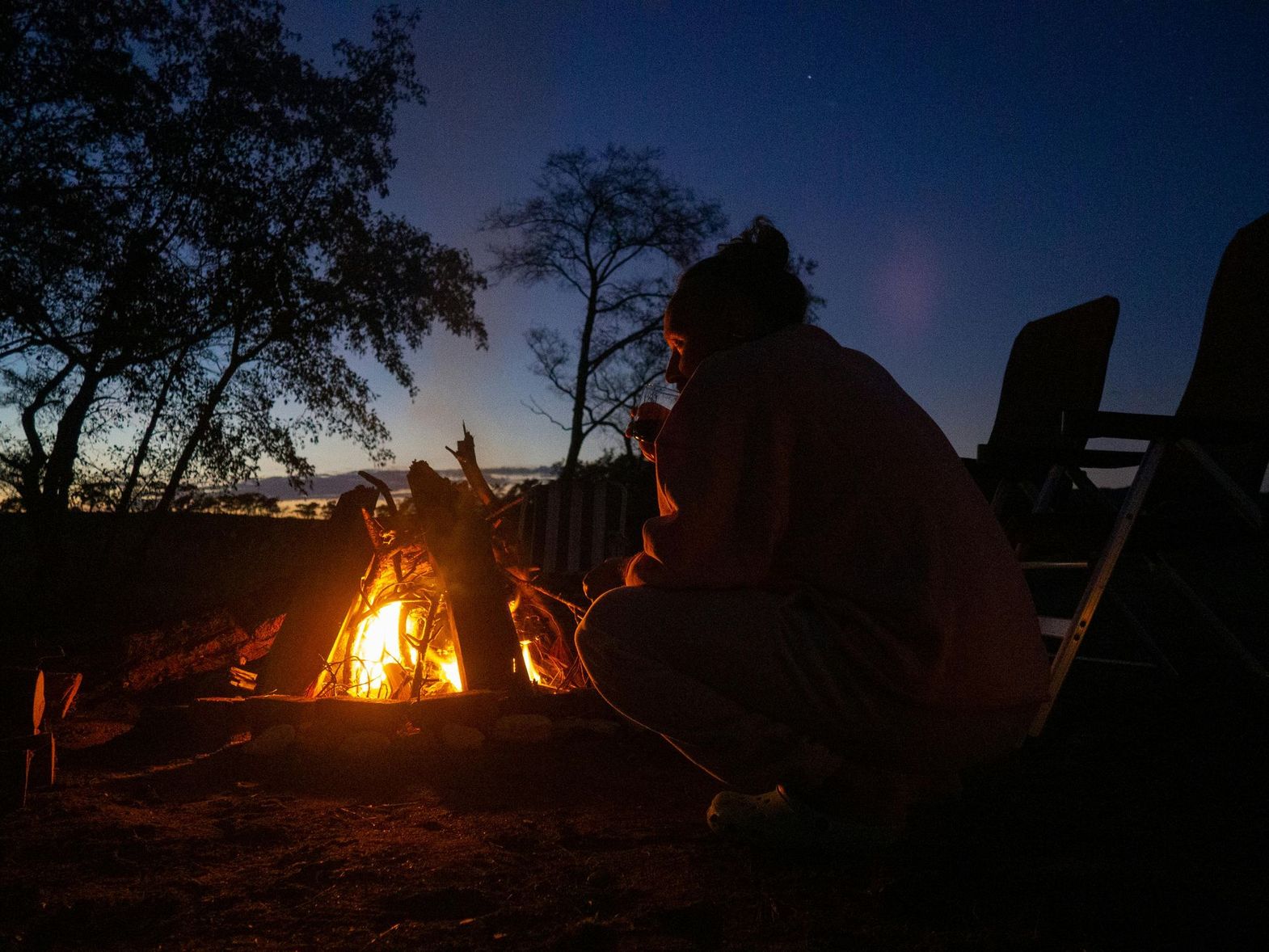 People sitting by a campfire at dusk in a wooded outdoor setting