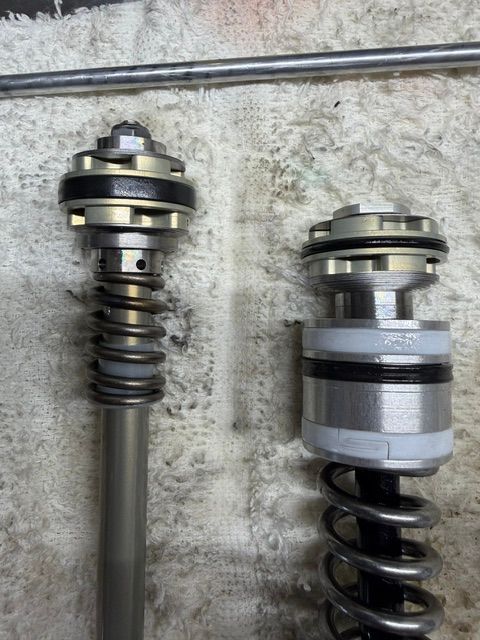Two metal suspension struts with coil springs, standing upright against a concrete wall.