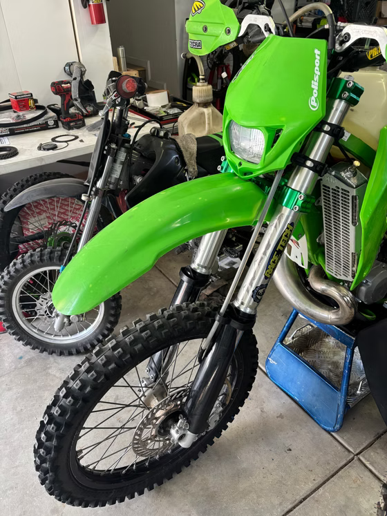 Bright green dirt bike in a garage, with other motorcycles and tools in the background