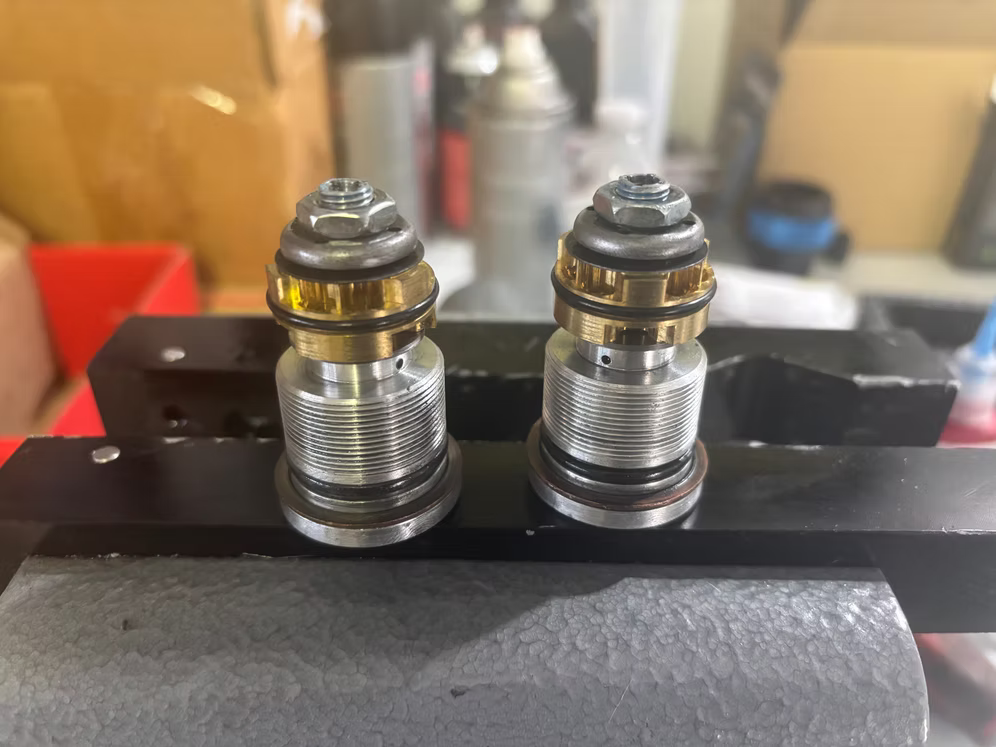 Two metal spark plugs mounted on a black engine part, with a blurred workshop background