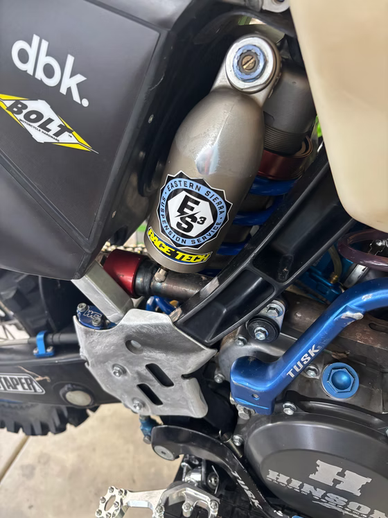 Close-up of a dirt bike engine and suspension with blue and silver parts, covered in stickers.