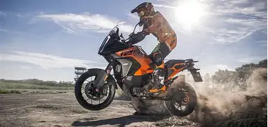 Motorcyclist riding an orange adventure bike through dust on a dirt trail at sunset
