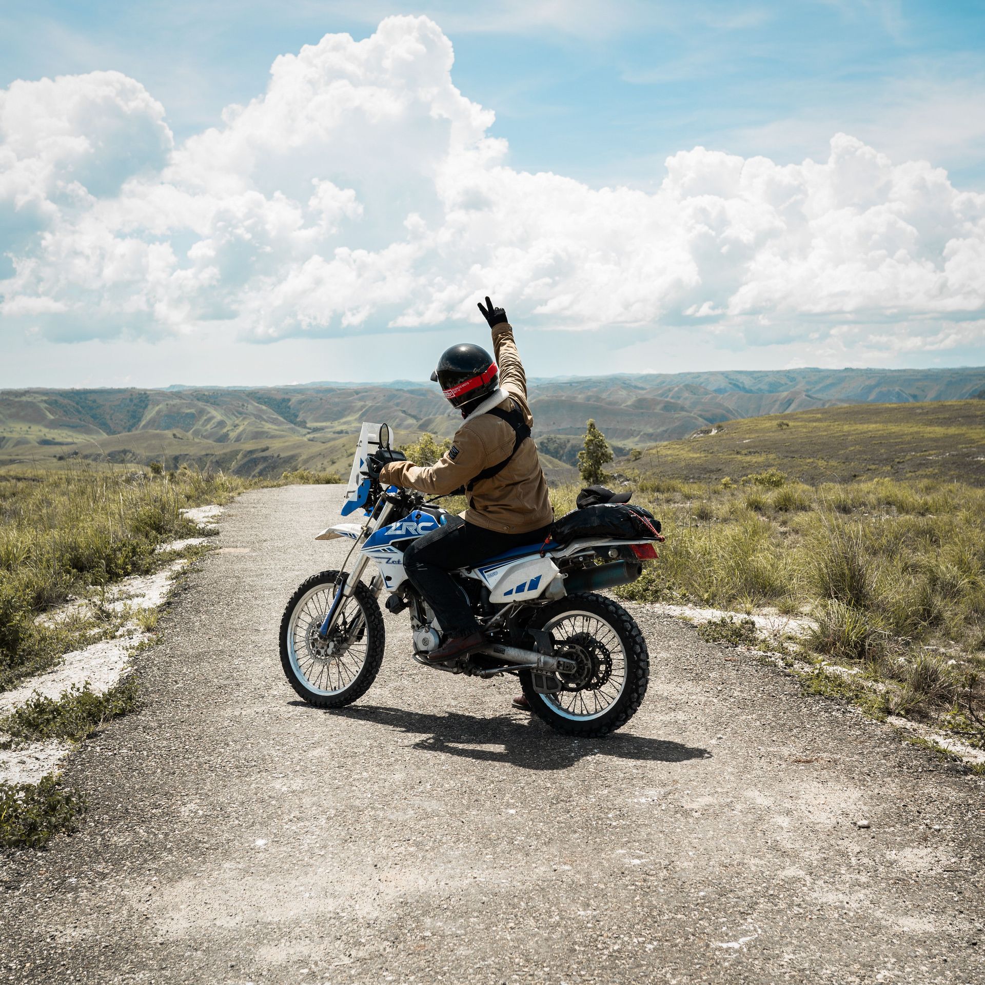 Motorcyclist waving on a dirt road in a grassy mountain landscape under a bright cloudy sky