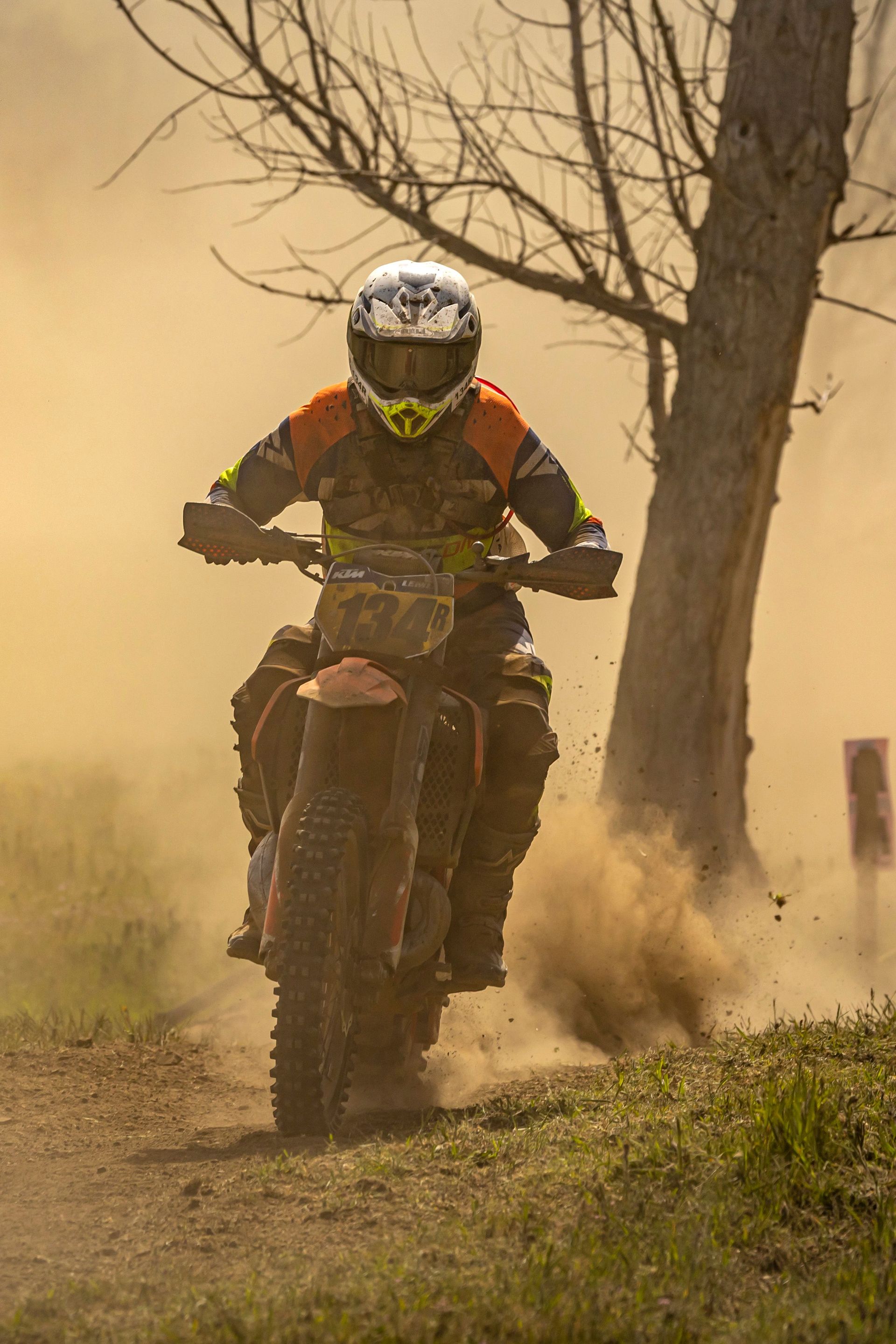 Motocross rider in neon gear racing through dust on a dirt trail near a tree