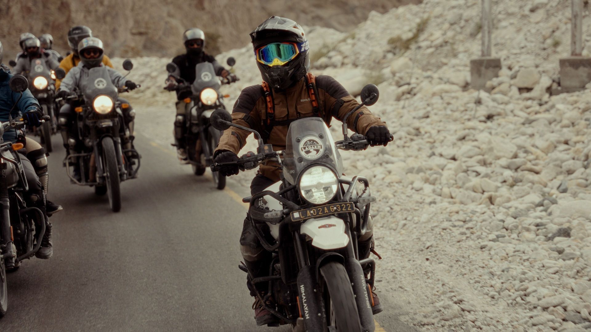 Motorcyclists ride through a rocky mountain road, headlights on, in dusty conditions