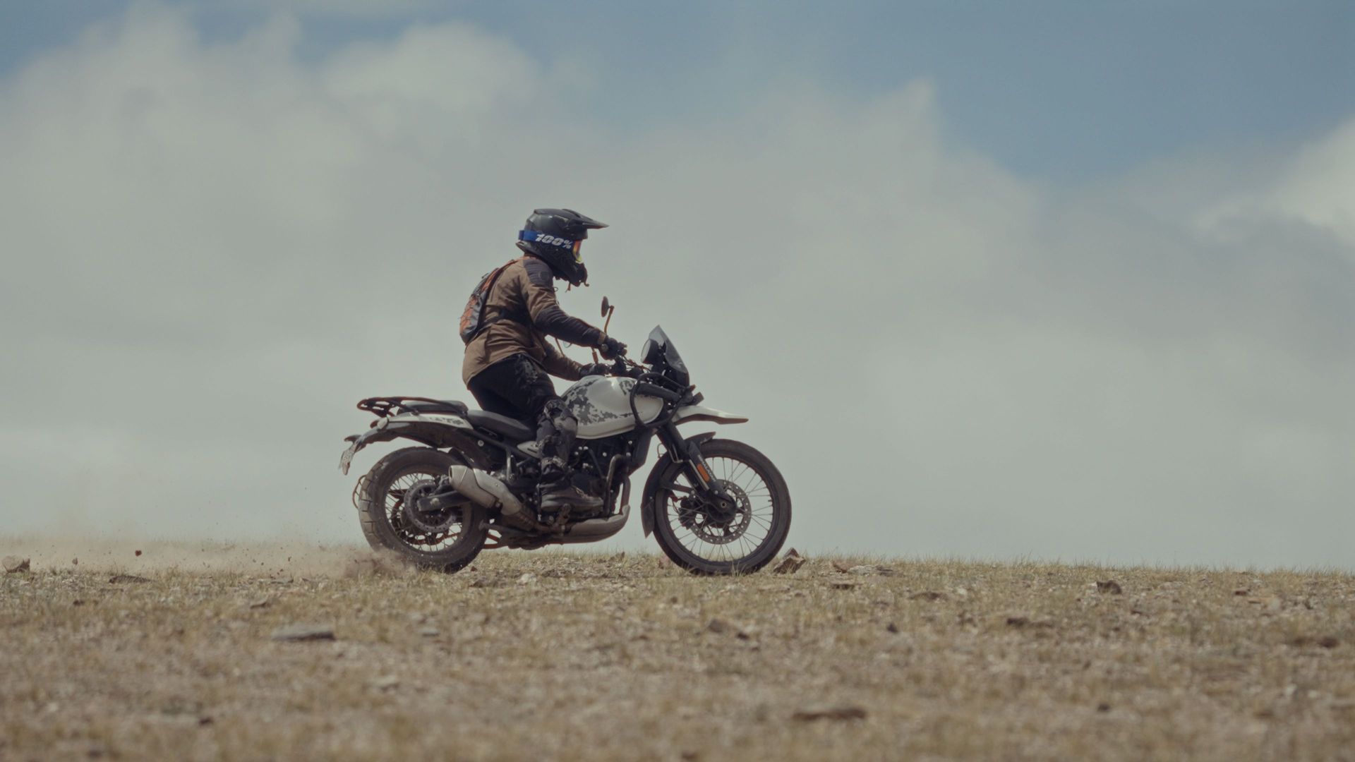 Motorcyclist riding across a dusty open plain under a cloudy sky