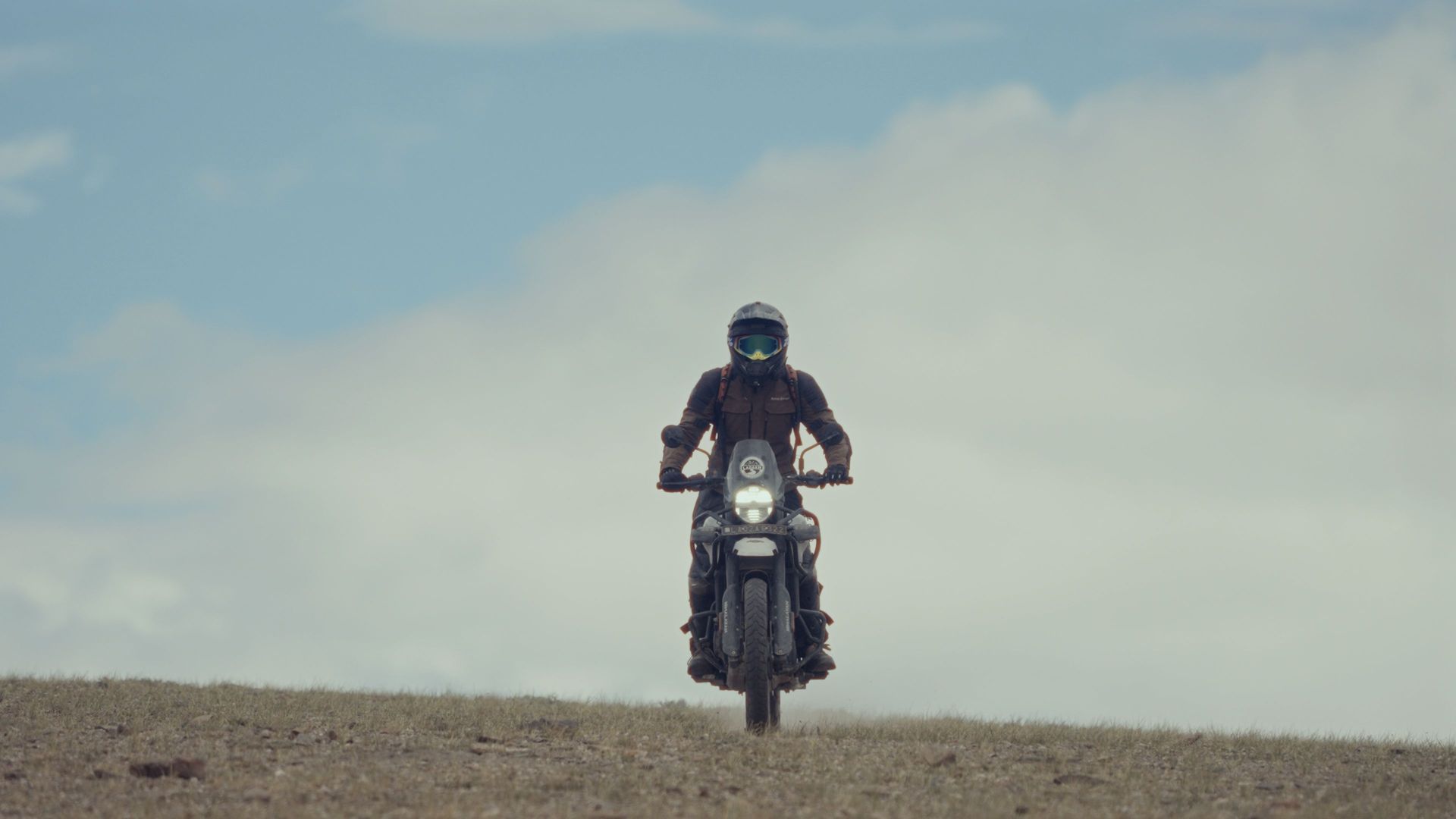 Motorcyclist riding toward the camera on a dirt ridge under a cloudy sky