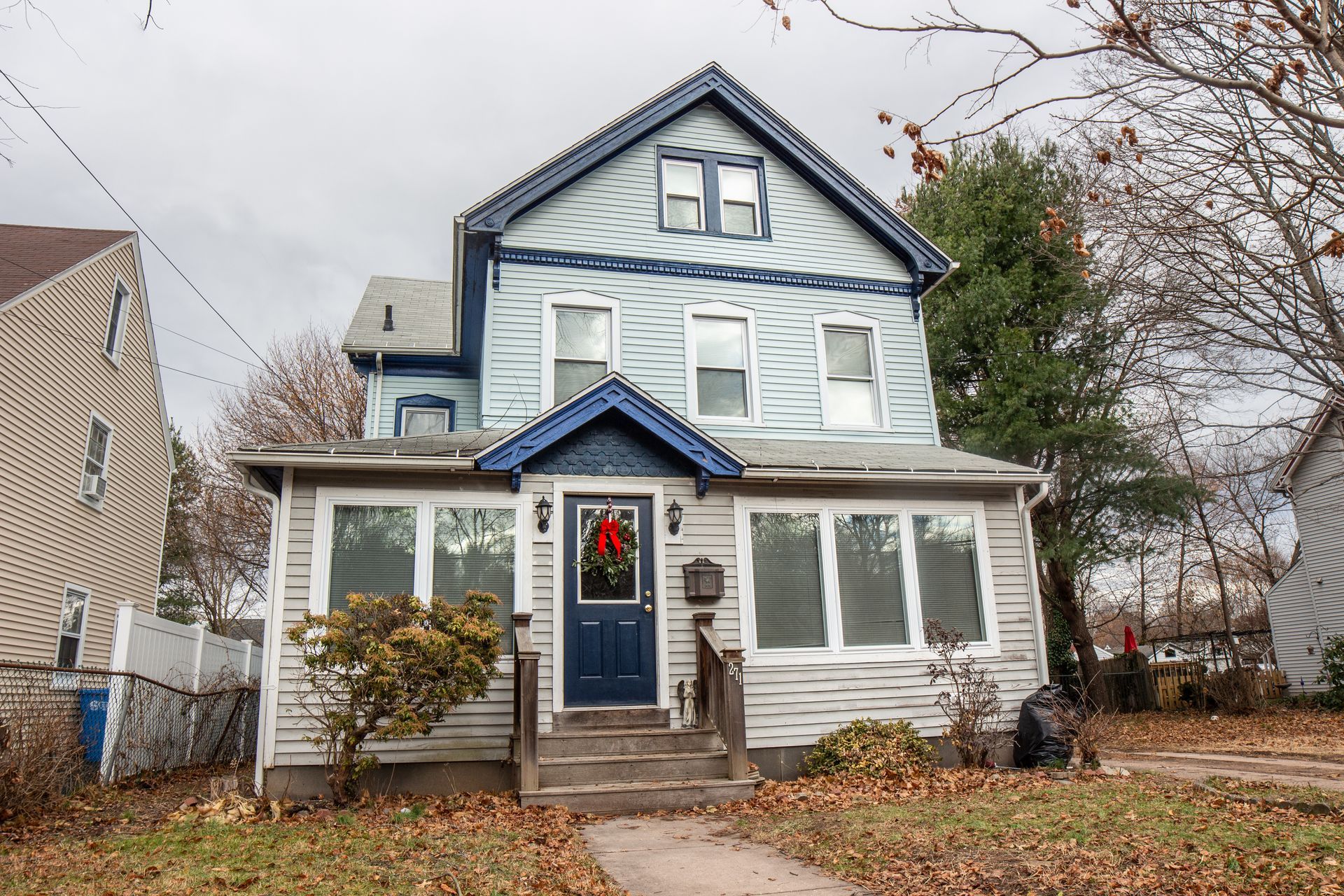 A two-story blue and grey house with a gabled roof, a front porch, and a blue front door decorated for the holidays.