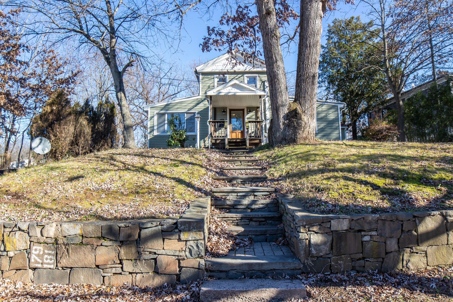 A green house atop a grassy hill with stone retaining walls and steps leading to the front porch on a sunny day.