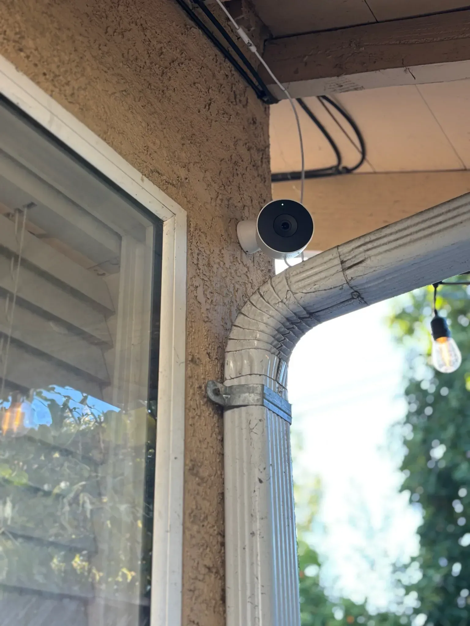 A round, white security camera mounted on a textured exterior wall next to a window and a white metal downspout.