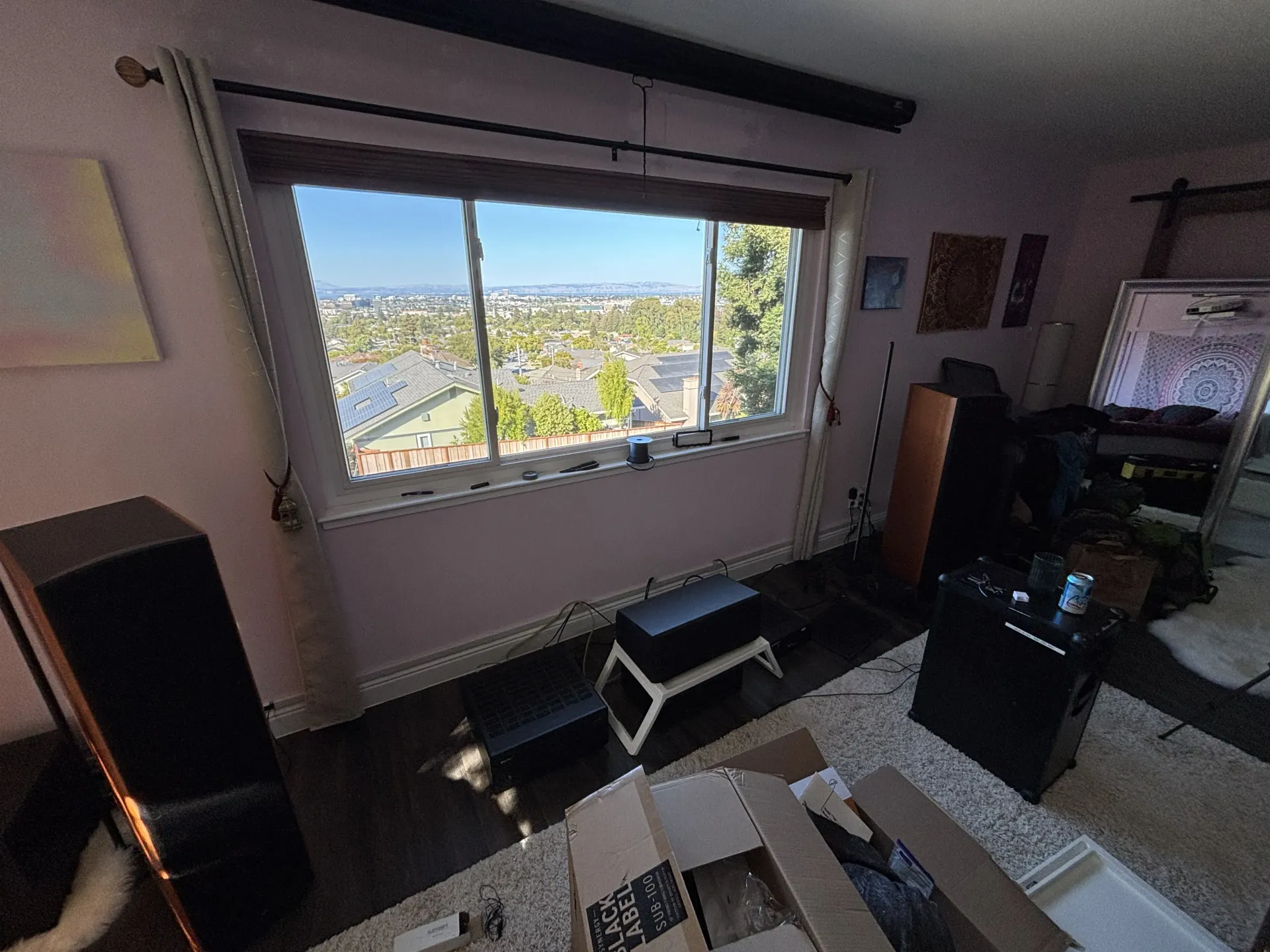 A view of a living room with large speakers, a center channel, an open shipping box, and a window overlooking a cityscape.