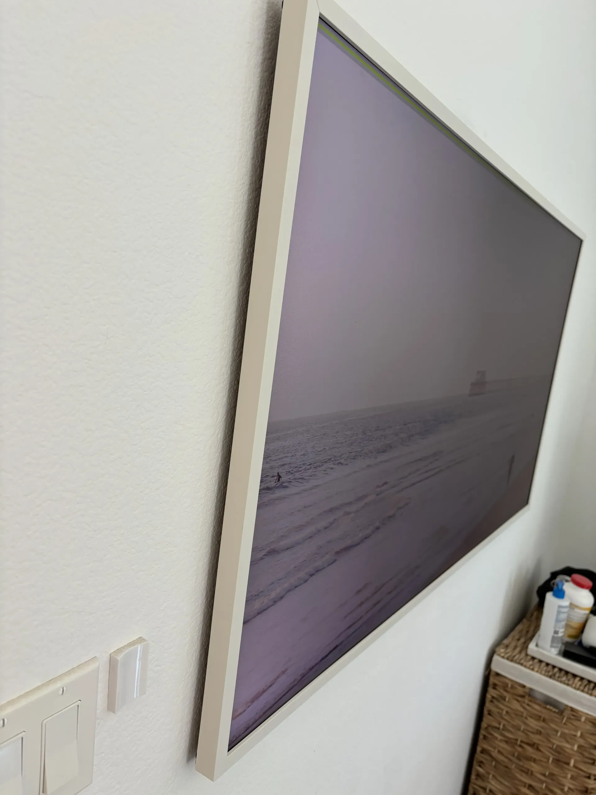 A framed, purple-toned landscape art print hanging on a white wall above a wicker basket.