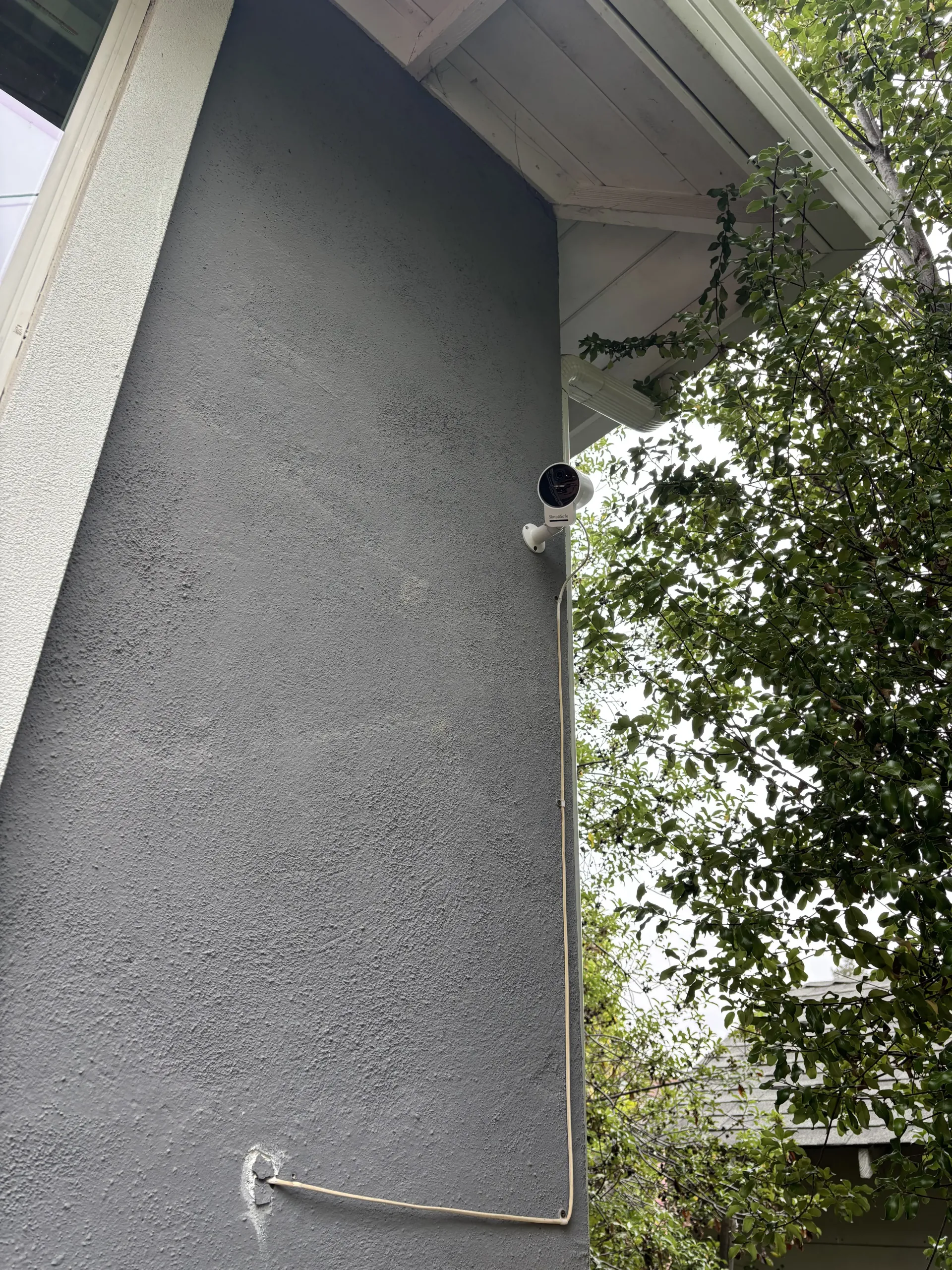 A security camera mounted on a textured gray exterior wall with a visible white cable running down to a wall entry point.