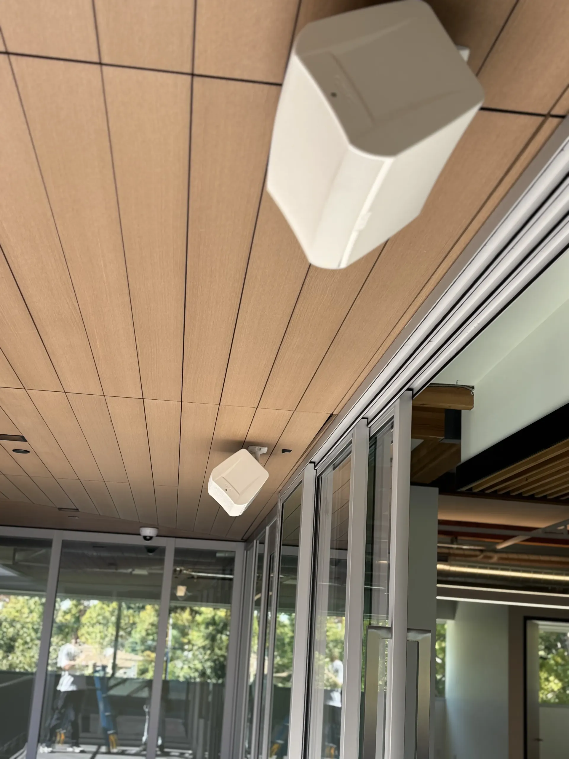 Two white speakers mounted to a wooden-slatted ceiling near a wall of vertical glass panels and window blinds.