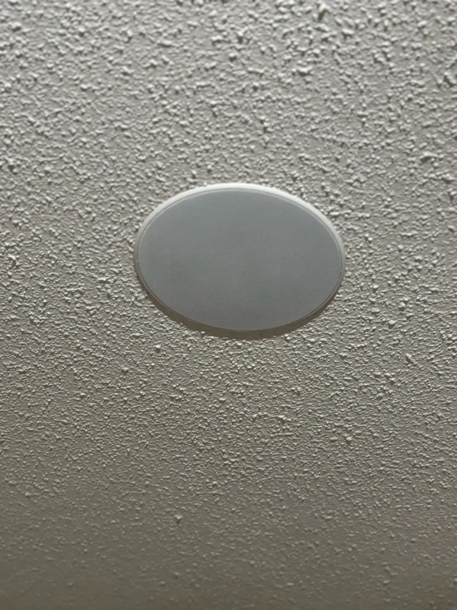 A round, white recessed ceiling speaker with a mesh grille mounted in a textured, off-white ceiling.