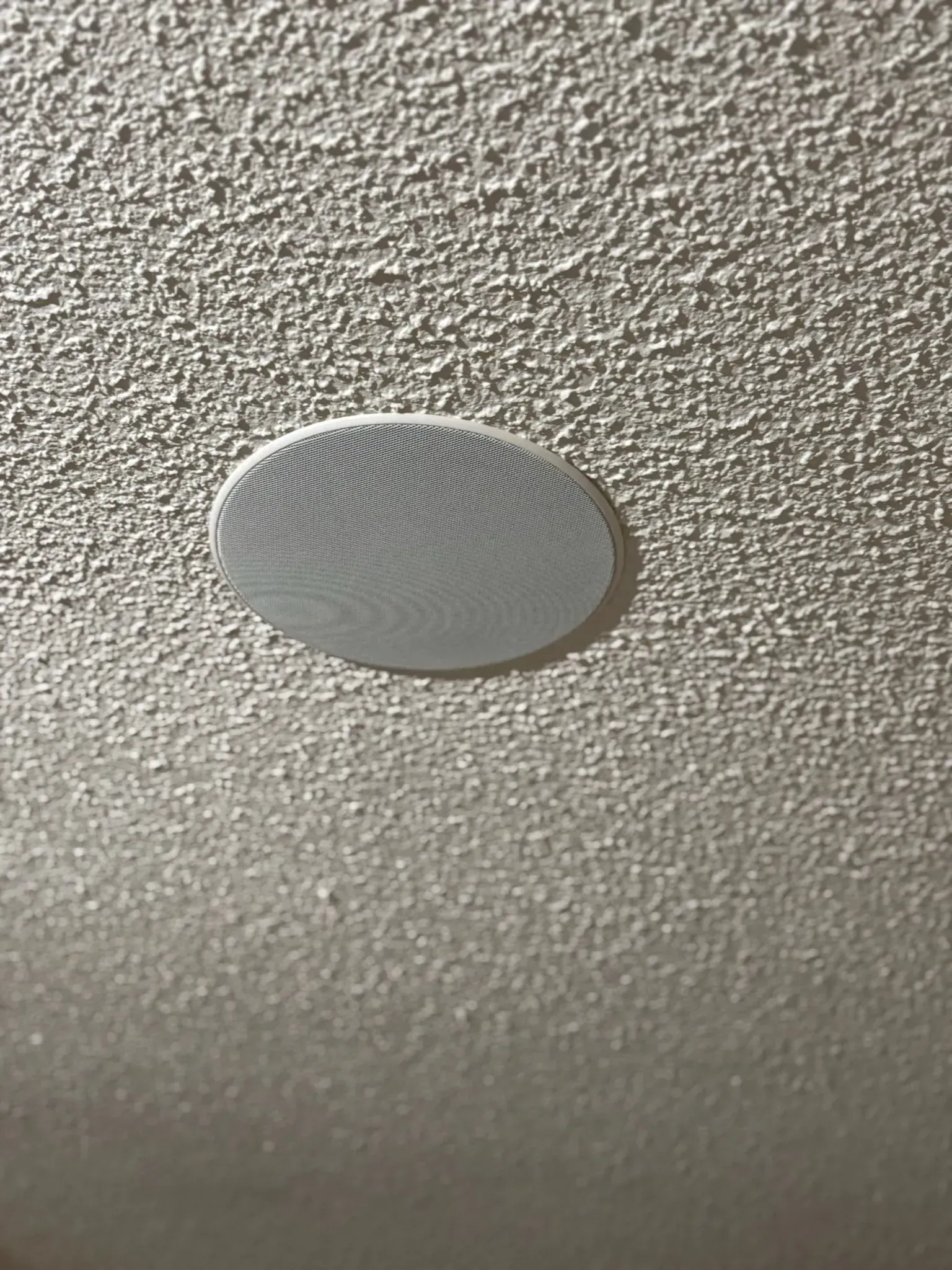 A circular ceiling-mounted speaker with a textured white grill installed in a popcorn-textured ceiling.