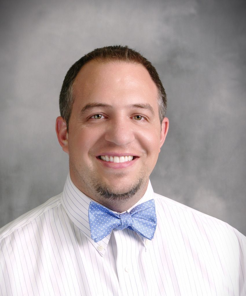 Smiling person in a white shirt and light blue bow tie against a gray studio backdrop