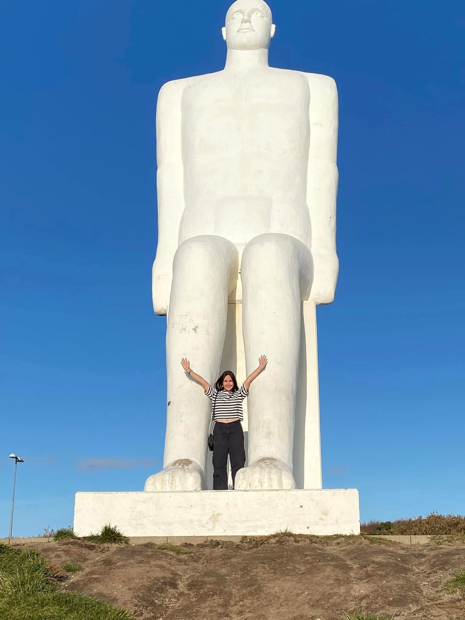 En kvinde står foran en massiv hvid statue af en mand med armene hævet mod en blå himmel.