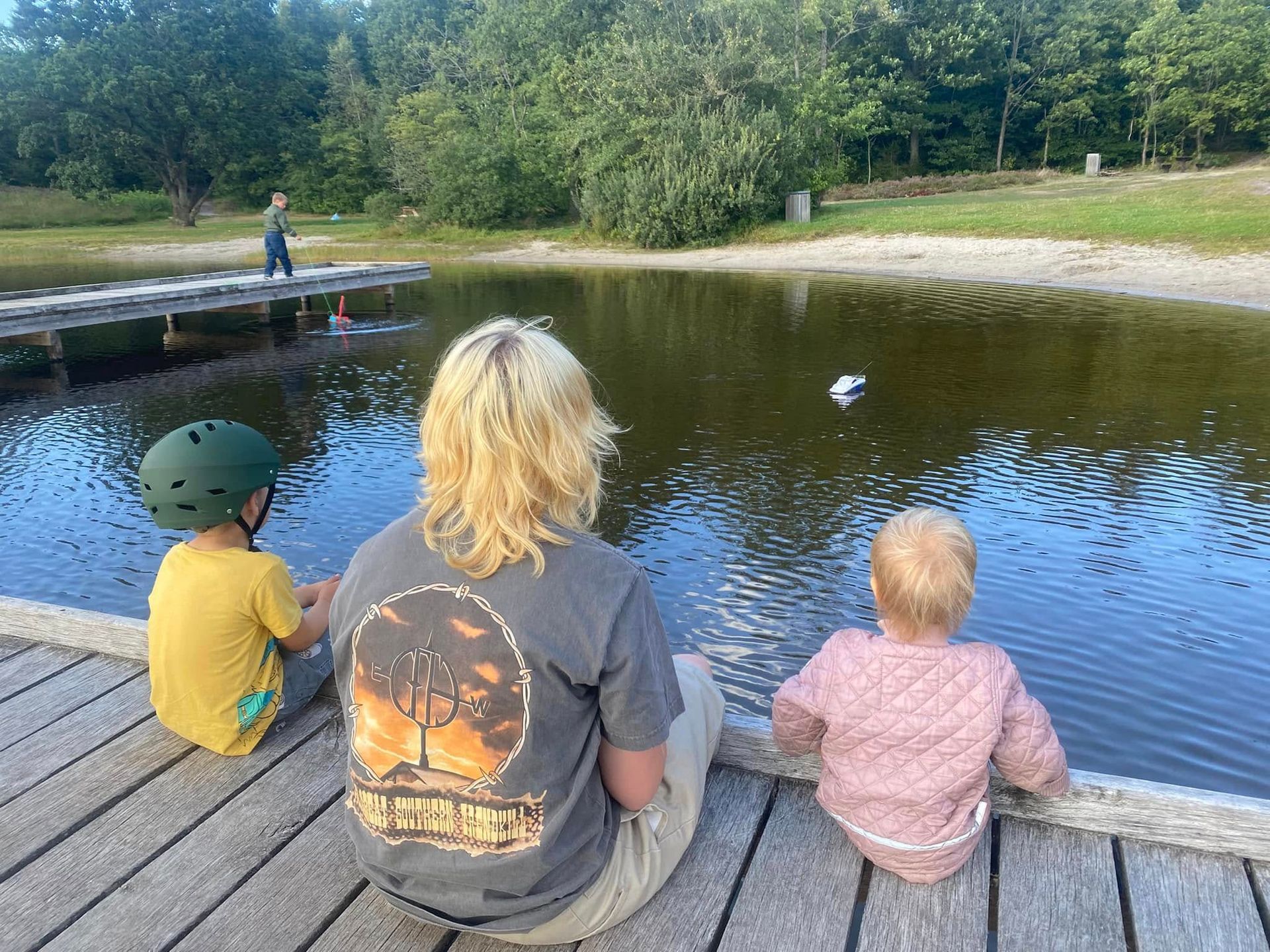 En kvinde og to børn sidder på en træbro og kigger på en damm. En dreng går på en mole i det fjerne.
