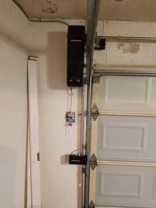 A black wall-mounted garage door opener and smaller electrical component installed on a white wall next to a garage door.