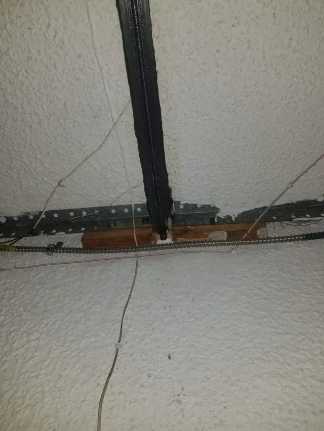 A black metal ceiling track intersects with a horizontal wood support block and metal mounting hardware on a textured wall.