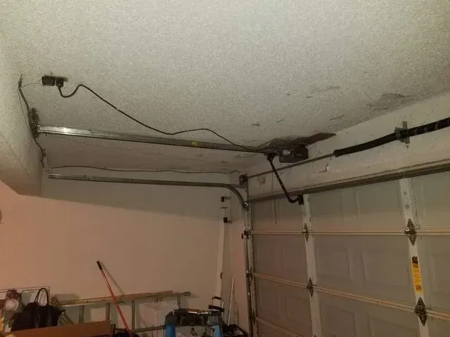 An interior view of a garage ceiling showing a garage door opener motor, power cord, and door tracks.