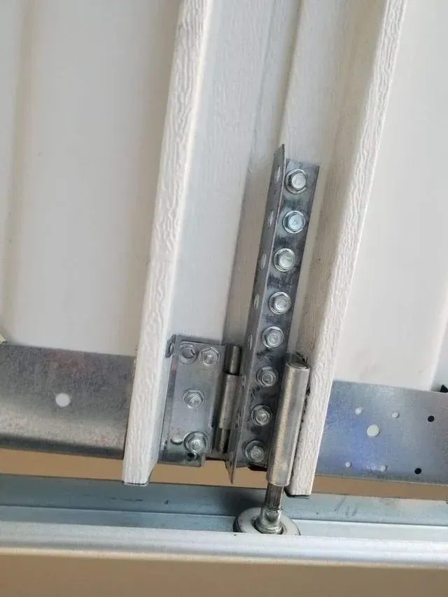 A metal hinge and roller assembly attached to the edge of a white garage door panel.