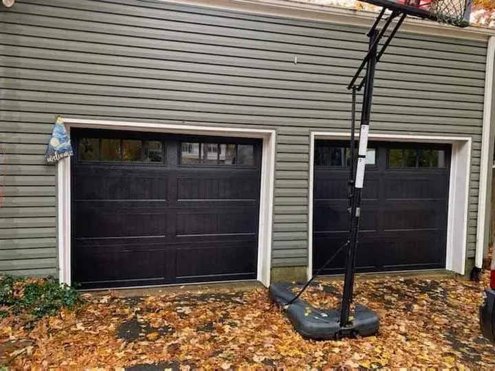 A residential gray garage with two black doors, featuring a basketball hoop stand on a driveway covered in autumn leaves.