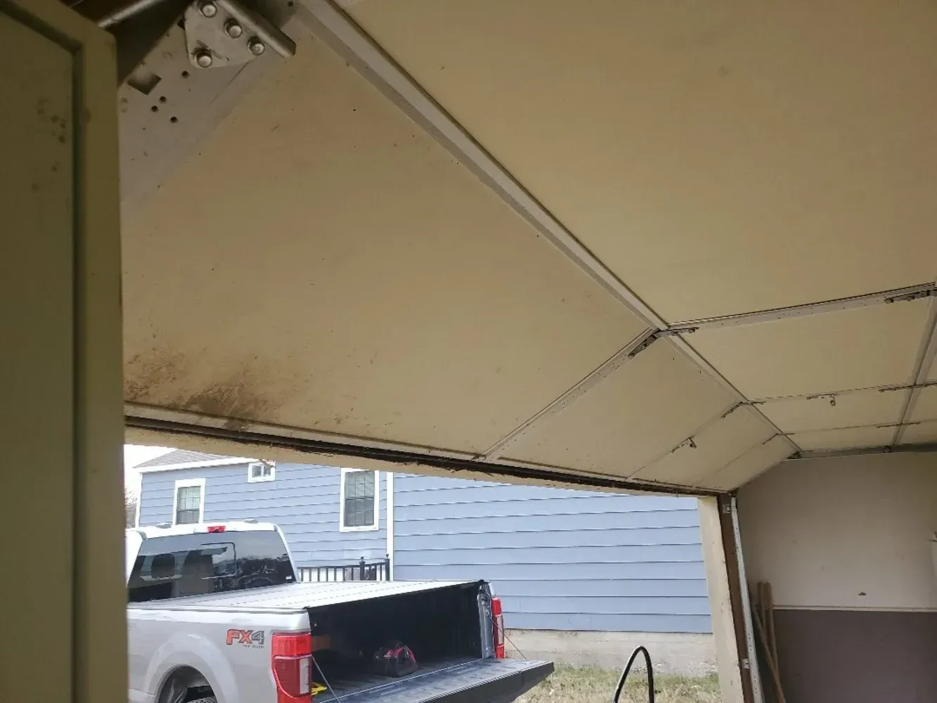 A view from inside a garage looking out at a white pickup truck parked in front of a blue-sided house.