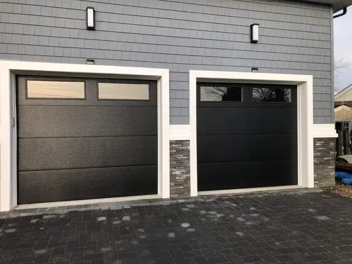 Two modern black garage doors with top windows, set in a house with grey shingle siding and stone accents.