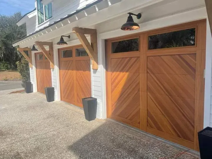 Three wooden garage doors with diagonal paneling and top windows, beneath black light fixtures on a white exterior wall.