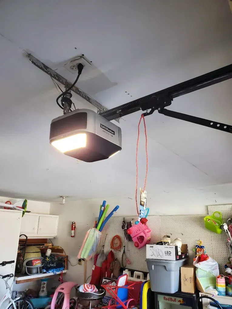 A garage door opener is mounted on the ceiling with an orange emergency release cord hanging down in a cluttered garage.