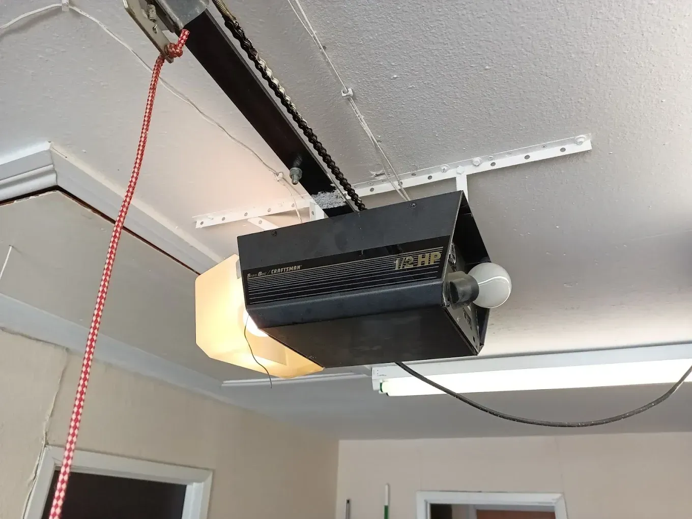 A black garage door opener with a light bulb mounted to a ceiling with metal support brackets and a red release cord.