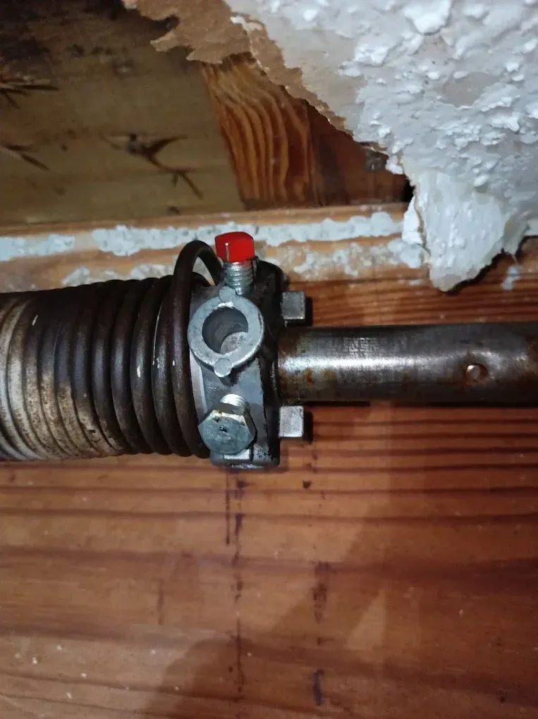 A close-up view of a metal torsion spring and a winding cone with a red cap mounted on a shaft against a wooden ceiling.
