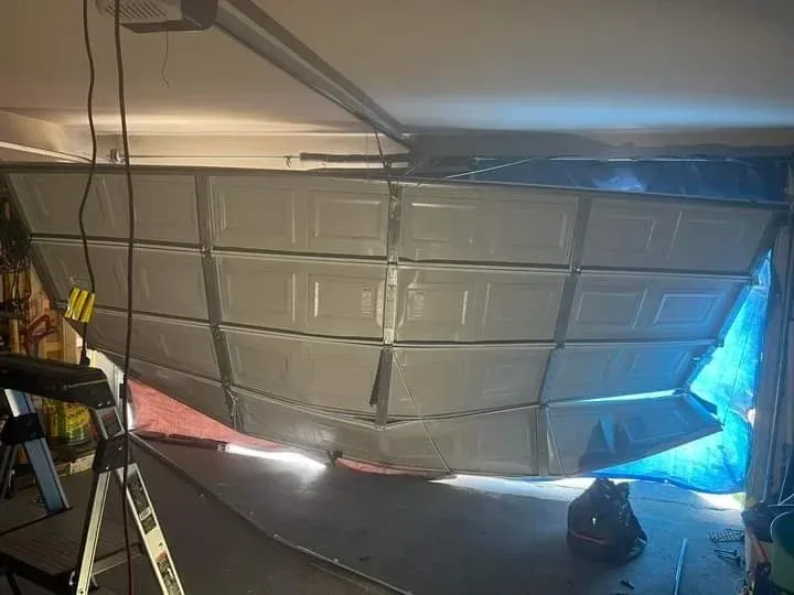 A broken, bent white garage door hanging at an angle from its tracks inside a dimly lit garage.