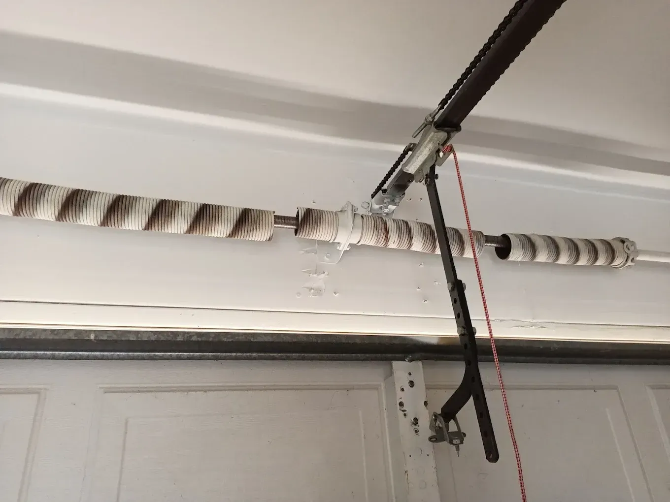 A broken metal torsion spring on a horizontal shaft above a white garage door, with a red emergency release cord hanging.