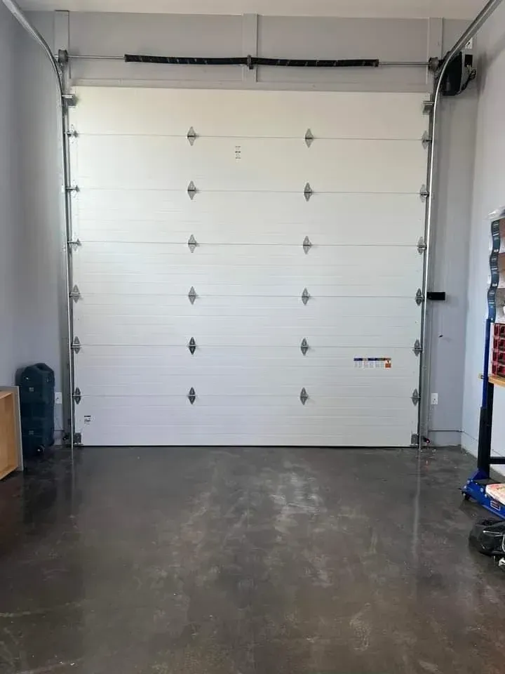 A white, multi-panel garage door closed inside a garage with grey walls and a dark concrete floor.