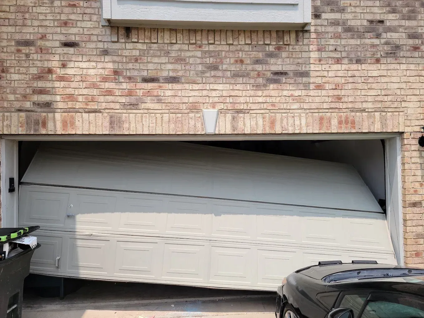 A beige, sectional garage door on a brick house is derailed, sagging crookedly in the frame.