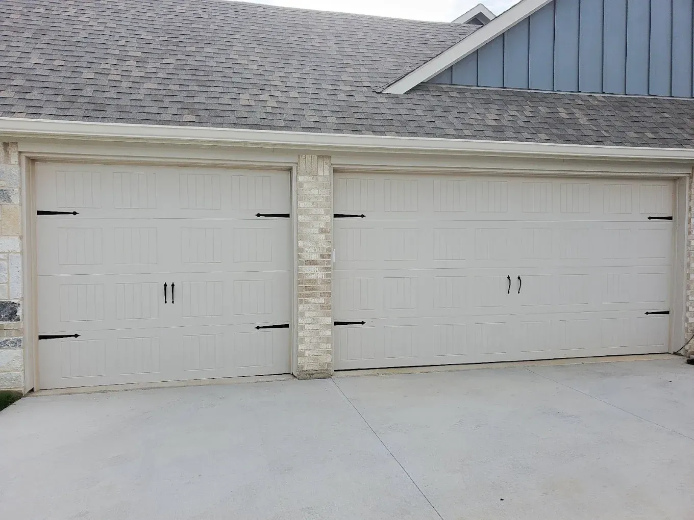 Two beige garage doors with black ornamental hinges and handles set against a stone-accented home exterior.