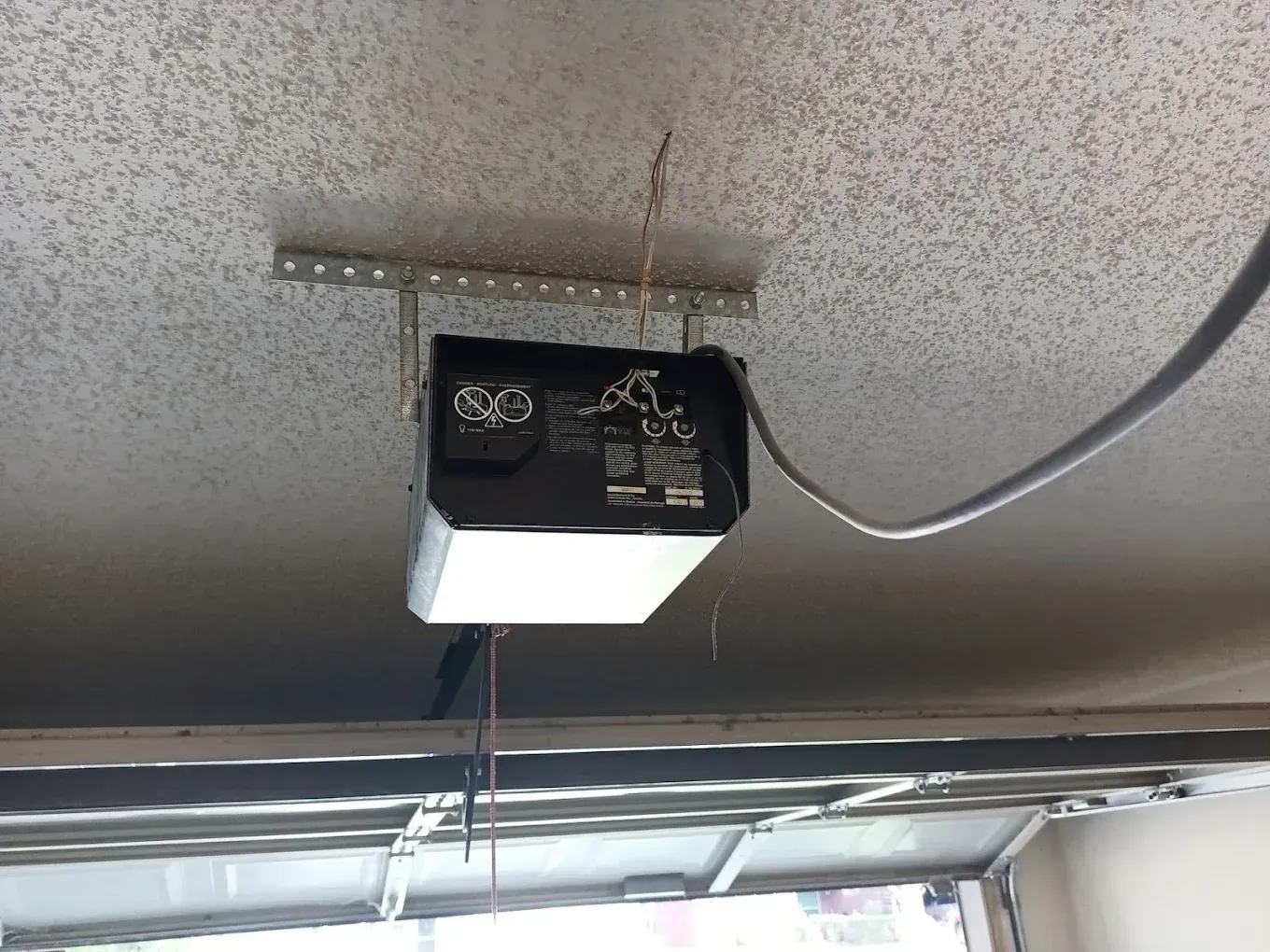 A black garage door opener motor hanging from a ceiling-mounted metal bracket with a power cord extending to the right.