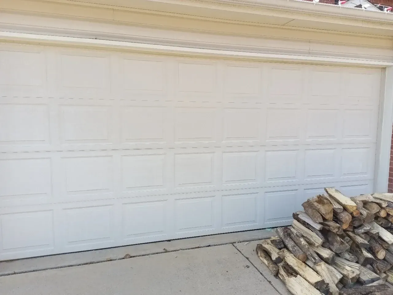A white, paneled garage door next to a large pile of firewood on a concrete driveway.