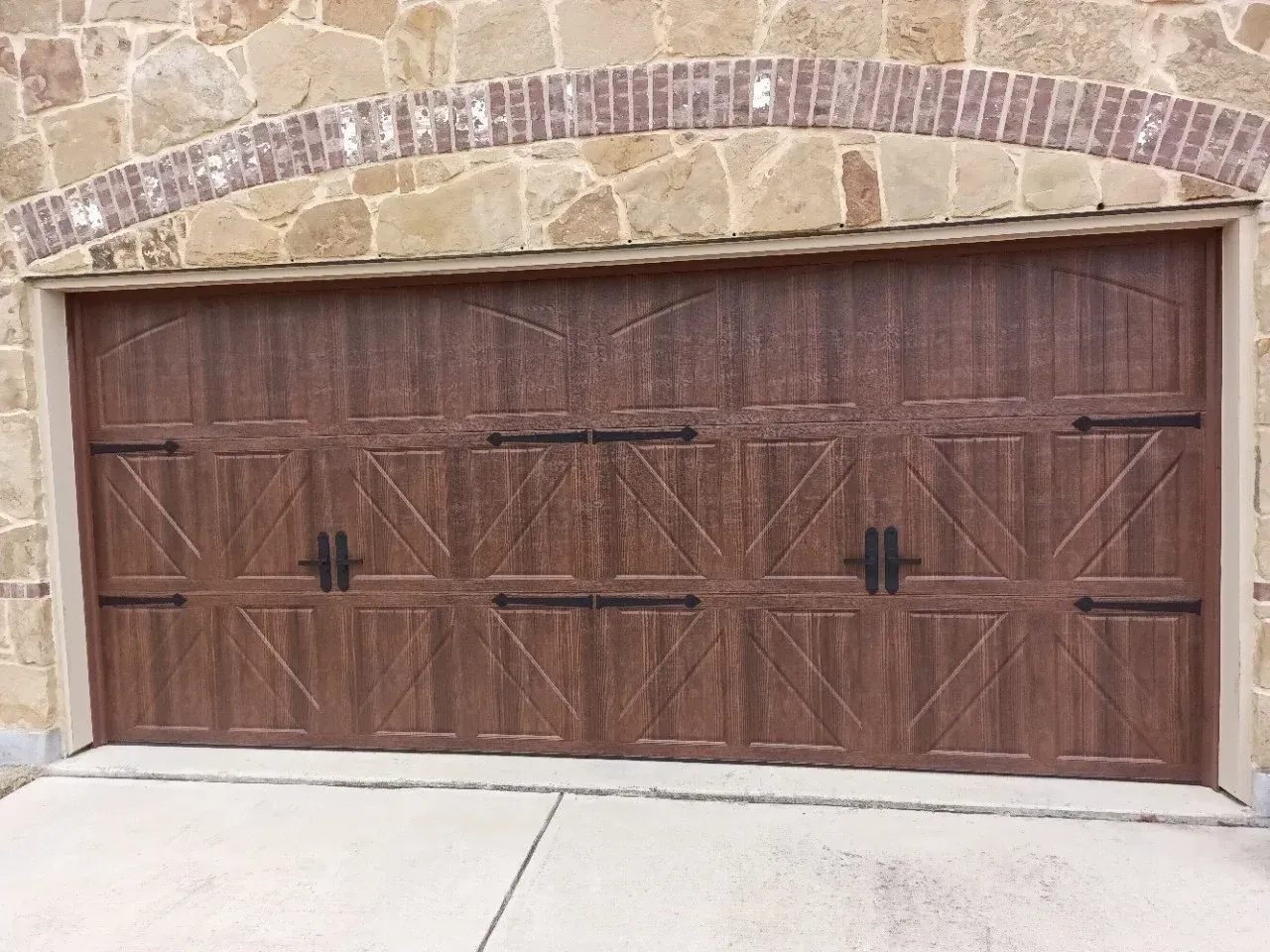 A brown wooden-style carriage garage door with black decorative hinges and handles, set under a brick and stone archway.