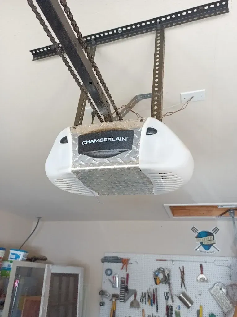 A white Chamberlain garage door opener hangs from the ceiling above a garage workshop with a pegboard tool organizer.
