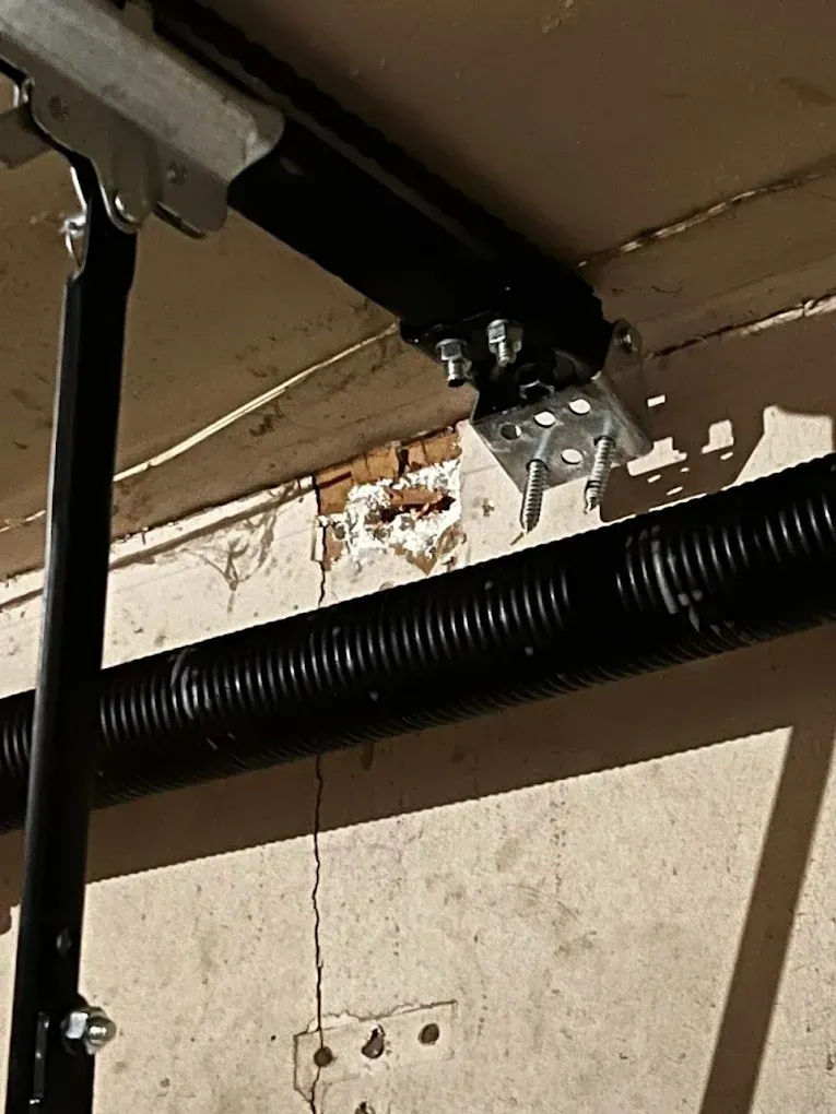 A black garage door opener rail bracket is mounted to a wall with a prominent vertical crack above the fastener.