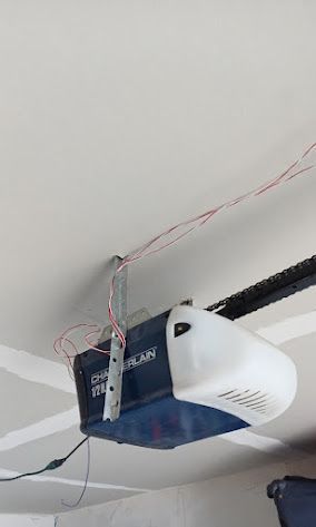 A white and blue Chamberlain garage door opener mounted to a ceiling with exposed, loose wires hanging nearby.