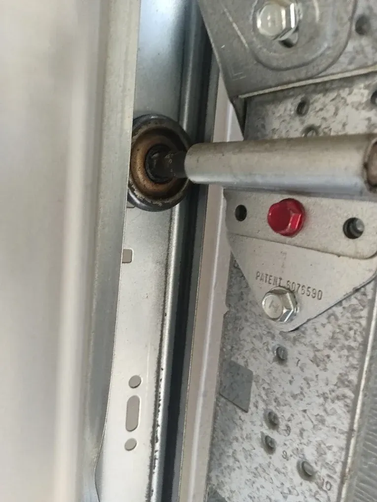 A garage door metal roller sits inside a vertical metal track, attached to a steel hinge plate with a red bolt.
