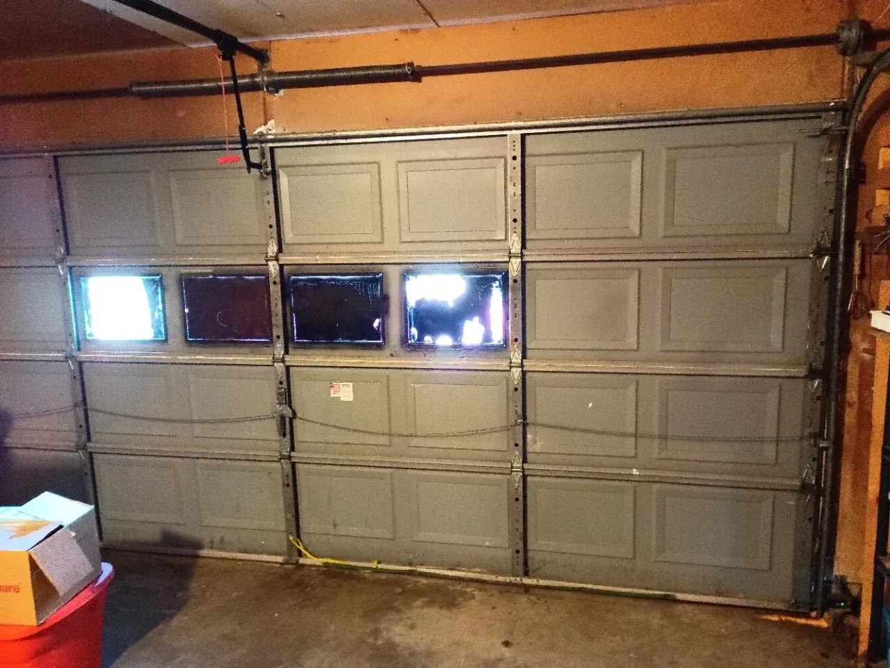 A grey sectional garage door with four windows in the second row, viewed from inside a dim garage.