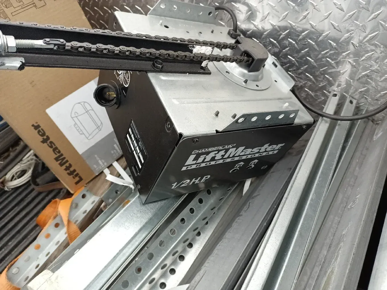 A LiftMaster garage door opener motor unit with a chain rail, resting on metal tracks in the bed of a truck.